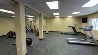 This fitness center offers a comprehensive workout environment designed to accommodate a range of exercise needs. The space is laid out across a single open floor, providing ample room to move freely between equipment zones. Structural columns are integrated throughout, dividing the area into functional sections without compromising the overall sense of openness.

The flooring consists of durable, speckled rubber or carpet tile material well-suited for high-traffic fitness use, while the drop-panel ceiling features evenly distributed recessed fluorescent lighting that keeps the entire space bright and well-illuminated. Warm beige walls with dark base trim contribute to a clean, neutral aesthetic throughout.

Cardio equipment is prominently featured and includes multiple treadmills, a stationary recumbent bike, an upright exercise bike, and an elliptical trainer. A dedicated strength training area is equipped with a multi-station cable and weight machine, offering users options for resistance-based workouts. A wall-mounted shelf or ballet barre along one wall suggests additional space for stretching or balance exercises. The facility is thoughtfully arranged to allow simultaneous use of both cardio and strength equipment, making it a versatile and functional space for fitness enthusiasts of all levels.