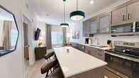 Located at 880 E Tucson Marketplace Blvd in Tucson, AZ, this thoughtfully designed one-bedroom, one-bathroom unit offers a cohesive modern aesthetic with warm neutral tones and light wood-style flooring throughout. The open-concept kitchen and living area serves as the heart of the unit, anchored by a central kitchen island with built-in shelving and seating, illuminated by distinctive teal pendant lights. The fully equipped kitchen features stainless steel appliances — including a refrigerator, range, microwave, and dishwasher — complemented by taupe textured cabinetry and clean white countertops. The living area provides a comfortable retreat with large windows dressed in layered blinds and drapes that invite natural light, alongside wall-mounted artwork with a Southwest-inspired color palette. The bedroom is a serene space with a wood-framed platform bed, dual nightstands, and a natural light-filled window with soft drapery, plus access to a walk-in closet. The bathroom includes a wide vanity with ample counter space, open shelving for storage, and a stacked in-unit washer and dryer — a highly practical feature. The space is efficiently laid out, making excellent use of every square foot with a clean, contemporary style.