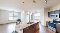 This well-appointed single-floor residence offers a thoughtfully designed open-concept layout that seamlessly connects the kitchen, dining, and living areas. The kitchen serves as the centerpiece of the space, featuring a large granite-topped island with a double-basin sink, dark espresso cabinetry, a full suite of stainless steel appliances — including a refrigerator, electric range, microwave, and dishwasher — and a tile backsplash. Pendant lighting and a track light fixture illuminate the space, while multiple windows allow abundant natural light to flow throughout.

The living and dining areas extend naturally from the kitchen, offering hardwood-style flooring and clean, neutral walls that create a bright and versatile backdrop. Additional rooms include bedrooms with a mix of plush carpet and hardwood-style flooring, each equipped with windows and ceiling fans for comfort. A well-sized closet with wire shelving provides ample storage.

The bathroom is finished with matching dark cabinetry, a granite vanity countertop, a large mirror, and a full tub-shower combination. A dedicated hallway with a flush-mount ceiling light connects the various areas of the unit, creating a logical and efficient flow throughout this clean, contemporary residence.
