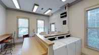 This shared laundry facility is designed with both functionality and comfort in mind. The room features a row of modern front-loading washers and dryers neatly arranged along one wall, providing ample capacity for residents. A generous laminate folding counter wraps along the center of the space, offering plenty of room for sorting and folding laundry. The neutral-toned walls are complemented by crown molding details near the ceiling, lending the room a polished, well-maintained appearance. Fluorescent ceiling fixtures provide consistent, bright overhead lighting throughout. Multiple windows fitted with horizontal blinds allow natural light to filter in, creating an inviting atmosphere with views of the surrounding greenery outside. A wall-mounted flat-screen television is positioned for easy viewing, adding a leisure element while residents wait. Seating is available near the windows, making the wait more comfortable. The space is clean, organized, and thoughtfully laid out to accommodate multiple users simultaneously, reflecting a practical and user-friendly design approach.