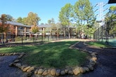 This outdoor recreational space features a well-maintained tennis court branded with the Thirty Cobb logo, finished in a classic blue and green hard-court surface. The court is fully enclosed by sturdy dark chain-link fencing, providing both safety and clear boundary definition for players. Overhead court lighting is mounted at the corners, allowing for extended use beyond daylight hours. The court is flanked by a natural landscape of mature pine and deciduous trees, which provide a pleasant wooded backdrop and shade along the perimeter pathway. Stone-lined borders demarcate the grounds, adding a structured yet natural aesthetic to the outdoor setting. The surrounding green lawn area offers open space adjacent to the court, complemented by well-kept landscaping throughout. In the background, a multi-story residential building is visible, reflecting the community-oriented nature of the facility. The entire area benefits from an open-sky setting that floods the space with natural light, creating an inviting and energetic atmosphere for tennis and outdoor activity.
