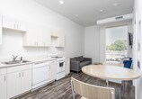 This single-floor unit in Clarksville, Tennessee offers a clean, minimalist design characterized by crisp white walls, wood-look vinyl plank flooring, and a straightforward, functional layout throughout.

The kitchen is efficiently arranged along one wall, featuring white cabinetry with ample upper and lower storage, a white corian-style countertop, a full-size refrigerator, an electric range, a dishwasher, and a microwave. The seamless cabinetry and neutral color palette give the kitchen a tidy, cohesive appearance. The kitchen opens directly into the living area, creating an open feel within the compact footprint.

The living area benefits from a large window fitted with horizontal blinds, allowing natural light to filter in while providing a view of the surrounding streetscape. The space is well-suited for a small seating arrangement.

The bathroom is divided into distinct areas, with a white vanity and toilet in one section and a separate walk-in shower stall featuring chrome fixtures in another. Wood-look flooring continues throughout the bathroom, maintaining a consistent aesthetic across the unit.

Overall, the space presents a clean, modern, and practical environment ideal for straightforward, comfortable living in the Clarksville area.
