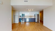 This contemporary single-floor residence showcases a clean, modern design with light hardwood flooring running throughout the space, creating a cohesive and airy feel. The open-concept layout connects the kitchen and living area seamlessly, maximizing the sense of space and flow.

The kitchen is a standout feature, equipped with full stainless steel appliances including a range, microwave, dishwasher, and refrigerator. Sleek, high-gloss white upper cabinetry contrasts elegantly with dark wood-toned lower cabinets, while a glass tile backsplash adds texture and visual interest. Light quartz countertops complete the polished look, and track lighting overhead ensures the space is well-lit.

The living area is open and versatile, benefiting from abundant natural light. The bedroom is generously proportioned, highlighted by floor-to-ceiling windows and a glass door that opens to a private balcony with expansive city views, flooding the room with natural light. A louvered closet door and additional storage are conveniently located near the entryway.

The bathroom features a wide vanity with dark wood cabinetry, a white countertop, a large mirror, and a walk-in shower. The unit's clean lines, neutral palette, and thoughtful layout make it a refined and functional living space.