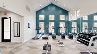 Located at 2323 Eldridge Parkway in Houston, TX, this fitness center is a thoughtfully designed workout facility that combines striking architectural elements with a full range of exercise equipment. The space is defined by its dramatic vaulted ceiling, which rises to a bold teal-painted peak accentuated by rows of multi-pane windows that flood the interior with natural light and offer views of the surrounding greenery. The crisp white walls contrast beautifully with the deep teal accent wall, creating an energizing yet clean atmosphere throughout. A geometric-patterned carpet in soft neutral tones covers the floor, adding visual interest while providing a comfortable surface underfoot. The cardio zone features multiple treadmills and elliptical trainers positioned along the windowed wall, while the strength training area is equipped with a cable machine, adjustable benches, a dumbbell rack, and various resistance machines. Full-length mirrors line one wall, enhancing the sense of openness and allowing users to monitor their form. Recessed lighting complements the natural light from above. Additional amenities include a water fountain station and a dedicated entry with a glass-panel door. The overall ambiance is bright, modern, and motivating, making it a functional and inviting space for fitness enthusiasts.