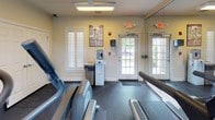 Located at 500 Carlen Ave in Lexington, South Carolina, this single-floor fitness center is a well-equipped facility designed to support a comprehensive workout experience. The space features rubber flooring throughout, providing a durable and safe surface for a wide range of exercises.

The room is outfitted with an array of cardio equipment, including multiple treadmills, elliptical trainers, and both upright and recumbent stationary bikes, offering users diverse options for aerobic training. A multi-function cable strength machine and a dedicated dumbbell rack round out the strength training offerings, making this a versatile facility for full-body workouts.

Large mirrored walls on one side of the room enhance the sense of openness and allow users to monitor their form during exercise. Natural light filters in through multiple plantation-shuttered windows, contributing to a bright and welcoming atmosphere. Ceiling fans and overhead fluorescent lighting ensure the space remains well-lit and comfortable. A wall-mounted television adds an entertainment option for those engaged in longer cardio sessions.

Fitness reference charts are posted throughout the room to guide users through various exercises. A water cooler is conveniently positioned near the exit, and glass-paned double doors provide direct outdoor access. The space is also equipped with safety features including visible exit signage and a security camera.