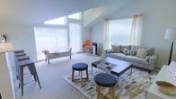 This single-floor space is defined by its soaring vaulted ceilings and an abundance of natural light streaming in through multiple large windows fitted with white horizontal blinds. The angled roofline adds architectural interest and contributes to the spacious, open feel of the interior. The main living area is thoughtfully arranged with a contemporary gray sofa accented by patterned throw pillows, a sleek rectangular coffee table with built-in compartments, and two round upholstered ottomans in charcoal gray. A geometric-patterned area rug in gold and white anchors the seating arrangement, adding warmth and texture to the neutral carpeted flooring. An accent chair in a vibrant orange provides a bold pop of color against the soft, light gray walls, while a classic upholstered bench near the window offers additional seating. Sheer patterned curtains frame one of the windows, softening the space and complementing the minimalist aesthetic. The overall ambiance is calm and welcoming, blending modern design elements with comfortable furnishings to create a versatile, light-filled environment suited for both relaxation and casual gatherings.