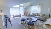 This single-floor space is defined by its soaring vaulted ceilings and an abundance of natural light streaming in through multiple large windows fitted with white horizontal blinds. The angled roofline adds architectural interest and contributes to the spacious, open feel of the interior. The main living area is thoughtfully arranged with a contemporary gray sofa accented by patterned throw pillows, a sleek rectangular coffee table with built-in compartments, and two round upholstered ottomans in charcoal gray. A geometric-patterned area rug in gold and white anchors the seating arrangement, adding warmth and texture to the neutral carpeted flooring. An accent chair in a vibrant orange provides a bold pop of color against the soft, light gray walls, while a classic upholstered bench near the window offers additional seating. Sheer patterned curtains frame one of the windows, softening the space and complementing the minimalist aesthetic. The overall ambiance is calm and welcoming, blending modern design elements with comfortable furnishings to create a versatile, light-filled environment suited for both relaxation and casual gatherings.