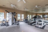 This single-floor fitness center is designed to offer a comprehensive workout environment in a bright, open setting. The main exercise area is generously sized, featuring wall-to-wall carpet flooring and warm, golden-toned walls that create an inviting atmosphere. Multiple ceiling fans are installed throughout the space, ensuring consistent air circulation during workouts.

The facility is well-equipped with a variety of cardio machines, including treadmills and elliptical trainers, as well as a multi-station strength training unit. A full-length wall mirror spans a significant portion of one side of the room, visually expanding the space and allowing users to monitor their form. Recessed lighting, combined with abundant natural light from multiple windows along the perimeter, keeps the space bright and airy throughout the day.

A dedicated open area provides ample room for stretching or floor exercises, and a wall-mounted television along with a water cooler add to the convenience of the facility. Fitness center rules are posted on the wall, indicating a shared, community-oriented space. Large glass French doors open directly to an outdoor area, offering refreshing views of the surrounding grounds. Decorative indoor plants are thoughtfully placed near the entrance, adding a touch of greenery to the functional environment. Security cameras are also present throughout the space.