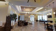 This single-floor space is designed with an open, airy layout that combines comfort and sophistication. The centerpiece of the main lounge area is a striking multi-tiered tray ceiling adorned with recessed lighting, crown molding, and a ceiling fan, creating a layered architectural focal point. A decorative chandelier adds an elegant touch, casting warm ambient light throughout the space.

The flooring throughout is rich hardwood, lending a warm and cohesive aesthetic that complements the earthy, golden-toned walls. The lounge area is furnished with plush seating arrangements, including dark leather sofas and accent chairs, anchored by a rustic wooden coffee table. A stone-accented fireplace along one wall adds a cozy, inviting element to the seating area, while tall indoor plants bring a touch of natural greenery to the interior.

Natural light filters in through multiple windows and a glass-paneled entry door, brightening the open floor plan. Adjacent to the lounge, a billiards table defines a dedicated recreation zone, making this space well-suited for both relaxed gatherings and social entertainment.