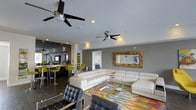 This single-floor space showcases a thoughtfully designed open-concept layout that seamlessly connects the living and bar or kitchen areas. The living space is anchored by a large white sectional sofa paired with a sleek glass coffee table, set atop a vivid multicolored area rug that adds a bold, energetic focal point to the room. Complementing the seating arrangement are black leather accent chairs, lending a sophisticated contrast to the lighter furnishings.

A decorative driftwood-framed mirror mounted on the gray-toned wall serves as a striking architectural accent, adding texture and visual interest. Two modern ceiling fans with dark blades are installed overhead, providing both functional airflow and a contemporary design element under the smooth white ceiling equipped with recessed lighting.

Adjacent to the living area, the bar or kitchen zone features vibrant lime-green bar stools positioned at a dark counter, creating a lively, social atmosphere. The overall ambiance of the space is modern and stylish, with a carefully curated color palette of neutral grays and whites energized by bold pops of yellow-green and multicolored accents throughout.
