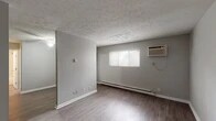 This single-floor unit is designed for functional, comfortable living and showcases a thoughtful combination of updated finishes and practical features throughout. The open living area is a standout space, featuring warm wood-look laminate flooring, neutral gray walls, and a decorative textured ceiling that adds visual interest overhead. A wall-mounted air conditioning unit and baseboard heating ensure year-round climate comfort, while a window allows natural light to filter into the room. An adjacent area provides additional living or dining space, also finished with the same consistent flooring and neutral palette, with a ceiling fan adding to the comfort of the space. The kitchen is well-appointed with slate-gray shaker-style cabinetry, dark countertops, a stainless steel sink, and black appliances including a refrigerator and an electric range with overhead ventilation. The cabinetry provides ample storage, and the layout makes efficient use of the available space. The bathroom is a refined highlight of the unit, featuring a tub-shower combination adorned with elegant marble-look tile surround, complemented by brushed nickel fixtures, a white vanity, and wood-look tile flooring. Together, these spaces create a cohesive, move-in-ready unit with modern updates and a clean, neutral aesthetic throughout.