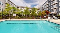 Located at 700 Bloomfield Ave in Bloomfield, Connecticut, this expansive outdoor amenity courtyard is nestled within a modern multi-story residential community. At its centerpiece is a generously sized swimming pool with crystal-clear turquoise water, accessible via built-in entry steps and pool-side railings. The surrounding deck is finished in smooth concrete, providing ample space for relaxation and recreation.

The sun deck is well-appointed with a generous arrangement of cushioned chaise lounge chairs and accent pillows, complemented by multiple large green market umbrellas that offer shaded retreats throughout the day. A charming wood-framed pergola with string lights anchors one corner of the courtyard, creating an inviting social gathering spot with additional seating.

Lush mature trees and carefully maintained landscaping add greenery and a natural buffer along the perimeter, enclosed by a sleek black iron fence that ensures both security and an open, airy feel. Colorful potted flowers and planters add vibrant seasonal accents throughout the space.

The courtyard is framed on multiple sides by the multi-story residential building, featuring modern gray and blue siding, large windows, and private balconies — creating a cohesive and sheltered environment. This outdoor space is designed to encourage community, leisure, and enjoyment in a well-maintained setting.