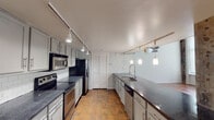 This two-floor space embodies an industrial loft aesthetic, blending raw architectural character with functional living areas. The main level features warm terracotta tile flooring throughout, anchoring the open layout with a grounded, earthy tone. Exposed concrete columns and large-scale silver HVAC ductwork running along the ceiling reinforce the industrial character, while a sleek white metal staircase with railing connects the two floors and serves as a visual focal point in the open living area. Large windows fitted with roller shades allow diffused natural light to fill the space. The kitchen is well-appointed and galley-style, featuring grey cabinetry, dark granite countertops, a tile backsplash, stainless steel appliances including a range, microwave, and dishwasher, and a double sink island illuminated by pendant lighting on a track rail system. A full bathroom on the main level includes terracotta tile flooring, a granite-topped vanity, and a tub-shower combination with large-format grey tile surround. The upper level features rich dark hardwood flooring, an open loft layout with metal railing overlooking the floor below, and exposed ductwork and raw ceiling details that carry the building's industrial heritage throughout both floors.