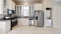 This single-floor residence offers a thoughtfully designed layout with a clean, neutral aesthetic carried consistently throughout. The kitchen is a standout feature, showcasing white shaker-style cabinetry with glass-front upper panels, gray quartz-style countertops, and a full suite of stainless steel appliances including a gas range, over-the-range microwave, dishwasher, and a side-by-side refrigerator with ice and water dispenser. Light-toned tile flooring and a window fitted with plantation shutters contribute to a bright, airy atmosphere. A dedicated entry door in the kitchen area adds convenient access. The living areas continue the light flooring theme with wood-look tile, complemented by warm beige walls and crisp white trim throughout. Rooms are well-lit by generously sized windows dressed with white horizontal blinds, allowing ample natural light while maintaining privacy. The bathroom is finished with a pedestal sink, a mirrored medicine cabinet, chrome fixtures including a towel bar and toilet paper holder, and a combination tub and shower. Above the toilet, a white cabinet provides additional storage. The overall ambiance of this residence is clean, modern, and move-in ready, with a cohesive design language that creates a welcoming and functional living environment.