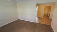 This two-story space presents a straightforward, functional layout with a mix of flooring materials that define distinct zones. The main living area is finished with wall-to-wall carpet in a warm brown tone, providing a comfortable underfoot feel. Adjacent to this carpeted zone, the flooring transitions to square tile in an earthy pattern, visible in the connecting hallway or secondary room, offering a practical surface for higher-traffic areas. The walls are painted in a soft, neutral cream tone, contributing to a clean and understated ambiance. White baseboards run consistently throughout, adding a crisp contrast to both the flooring and wall colors. HVAC vents are positioned along both the upper wall and lower floor level, indicating a dual-supply climate control system for efficient temperature management. The textured ceiling adds subtle visual depth to the interior. A doorway visible in the background connects the spaces and hints at additional rooms beyond, while a staircase is partially visible, leading to the second floor. Overall, the space offers a practical, neutral canvas suitable for a variety of uses.