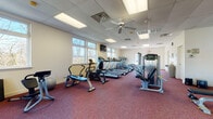This fitness center is a spacious, single-floor facility designed to accommodate a comprehensive workout experience. The room is finished with durable red speckled carpet flooring and features a drop ceiling with recessed fluorescent lighting panels and ceiling fans that promote air circulation throughout the space.

The cardio section is prominently equipped with multiple treadmills, elliptical trainers, and stationary bikes, arranged in organized rows along the windowed wall. Large windows line the perimeter, flooding the interior with natural light and offering views of the surrounding wooded landscape. A wall-mounted television is positioned near the windows for entertainment during workouts.

The strength training area includes a multi-station cable machine, a weight bench, and additional resistance equipment, providing users with a variety of options for resistance-based exercise. A stability ball and other accessories are also available within the space. A secondary area near the exit door houses additional elliptical machines and benefits from glass-panel doors that introduce further natural light and direct outdoor access.

The facility includes clearly marked emergency exits, a drop ceiling with ventilation, and neutral cream-colored walls that contribute to a clean, functional atmosphere. The overall layout is open and well-organized, allowing users to move freely between equipment zones.