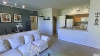 This single-floor residence features an open-concept layout that seamlessly connects the living area to the kitchen, creating a spacious and functional environment. The living room is finished with plush carpeting and neutral walls accented by a bold slate-blue feature wall, adding depth and visual interest to the space. A decorative floating shelf provides a subtle architectural detail, while crown molding frames the room with a clean, polished look.

The kitchen is positioned at the rear of the open floor plan and is equipped with a raised breakfast bar countertop, an undermount sink, a full-size refrigerator, and warm wood-toned cabinetry. Track lighting illuminates the kitchen workspace efficiently. A walk-in closet is accessible from the main living area, offering generous built-in storage with visible shelving and hanging space.

Ceiling-mounted vents and recessed lighting are distributed throughout, ensuring consistent climate control and illumination. The overall aesthetic is cohesive and understated, combining practical design with a comfortable, welcoming ambiance suited for everyday living.