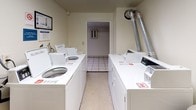 This shared laundry room is a functional, well-organized facility designed to serve multiple users. The room features light-colored walls and durable tile flooring throughout, creating a clean and practical environment. Two rows of white top-load washing machines and dryers line opposing walls, providing ample capacity for residents. A wall-mounted folding shelf offers a convenient surface for sorting or folding laundry, accompanied by a decorative potted monstera plant that adds a touch of warmth to the utilitarian space. Seating is available via a cushioned armchair positioned near the folding area, allowing users to wait comfortably. Safety is addressed with a wall-mounted fire extinguisher in a clearly marked cabinet. A waste bin is provided, and posted signage reminds users to keep the room clean and to refrain from disposing of household trash in the laundry room bins. Additional posted notices display operating hours of 8 AM to 10 PM. The room connects to an adjacent utility area visible through a secondary doorway, which houses an electrical panel and ventilation ductwork. Multiple electrical outlets are distributed along the walls for added convenience.