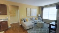 This single-floor residence features a thoughtfully designed living area that immediately draws attention with its warm yellow walls and rich hardwood-style flooring, creating a bright and inviting atmosphere. The main living space is generously proportioned, accommodating a large L-shaped sectional sofa dressed with patterned and solid accent pillows in complementary tones of grey, white, and gold. A triptych of abstract artwork in soft gold and cream hues is displayed prominently on the main wall, adding a sophisticated decorative focal point. A round upholstered ottoman serves as a functional centerpiece atop a grey geometric-patterned area rug, tying the seating arrangement together cohesively. Natural light filters in through a window fitted with layered curtains in charcoal and sheer tones, brightening the space without overwhelming it. A transitional hallway visible from the living area hints at additional rooms beyond, while wall-mounted art and a coordinating lamp contribute to the polished, unified aesthetic. The overall ambiance is warm, contemporary, and well-coordinated throughout.