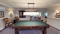 This single-story space is centered around a well-appointed billiards room, anchored by a substantial full-size pool table crafted from rich, dark wood with ornate carved detailing. The table's green felt surface is illuminated by a large pendant light fixture suspended from above, featuring a rustic natural wood accent piece paired with a wide, pleated fabric shade that directs focused light onto the playing surface. A wall-mounted cue rack holds an organized selection of billiards cues nearby, reinforcing the room's dedicated recreational purpose. The ceiling features white-painted tongue-and-groove paneling, contributing a clean, classic aesthetic throughout. White wainscoting lines the lower walls, adding a polished, traditional character. An open passageway leads directly into a comfortable lounge area beyond, where plush seating upholstered in coral and neutral tones creates a welcoming retreat. The lounge benefits from abundant natural light streaming through large windows, offering views of an outdoor setting. The overall ambiance is relaxed yet refined, blending a casual recreational atmosphere with thoughtful, classic design elements that make the space inviting and functional.