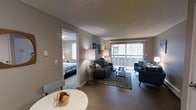 This single-floor residence is thoughtfully laid out to maximize functionality and comfort. The living area serves as the central hub of the space, featuring warm hardwood-style flooring that extends throughout, creating a cohesive and inviting atmosphere. A large sliding glass door fitted with vertical blinds dominates one wall, allowing natural light to filter in while offering access to an outdoor area. The living room accommodates a comfortable seating arrangement, including a tufted sofa and accent chairs centered around a coffee table atop a patterned area rug, making it well-suited for relaxing or entertaining.

A dining nook is positioned near the living area, furnished with a round white table and seating, adding a practical and casual dining option. Wall décor and coordinated floor lamps contribute to the overall warm ambiance. The textured ceiling adds a subtle architectural detail throughout.

Adjacent to the living space, the bedroom is accessible through a dedicated doorway and benefits from its own natural light source via a window. The room comfortably fits a full-sized bed and maintains the same neutral color palette seen throughout the rest of the space.
