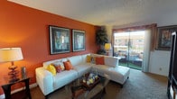 This single-floor space makes a striking first impression with its bold design choices and warm, inviting ambiance. The living area is the centerpiece of the space, anchored by a rich terracotta accent wall that adds depth and warmth to the room. A generously sized white sectional sofa, adorned with colorful throw pillows in shades of orange and yellow, provides ample seating and serves as a stylish focal point against the vivid backdrop.

A glass-topped coffee table with a dark wood frame sits at the heart of the seating arrangement, offering both functionality and an elegant visual contrast. Two large framed art pieces hang symmetrically on the accent wall, adding a curated, gallery-like quality to the space. A textured popcorn ceiling and neutral carpet flooring complement the overall warm color palette.

Natural light flows generously into the room through a full-length sliding glass door that opens onto a private balcony, where outdoor seating can be glimpsed alongside views of the surrounding exterior. Sheer curtains frame the doorway, softening the transition between indoors and out. A decorative floor lamp and lush indoor plant complete the room's inviting, lived-in character, making this space feel both comfortable and thoughtfully appointed.