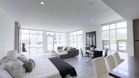 This contemporary single-floor residence offers a thoughtfully designed open-plan layout that effortlessly combines living, dining, and sleeping areas into one cohesive space. The interior is defined by an abundance of natural light, courtesy of expansive floor-to-ceiling windows that wrap around multiple sides of the space, framing views of the surrounding urban landscape and flooding the interior with daylight throughout the day.

The living area features a sleek, low-profile sectional sofa in neutral grey tones, complemented by a minimalist round coffee table that keeps the space feeling open and uncluttered. Adjacent to the living area, a compact dining zone is anchored by a glass-top table paired with contemporary black chairs, adding a refined, modern touch. A bold black-and-white figurative artwork on the wall provides a striking focal point and a sense of artistic character.

The sleeping area integrates seamlessly into the open layout, with a generously sized bed dressed in crisp white linens and a dark throw. The light hardwood-style flooring runs throughout, tying all zones together cohesively. Recessed ceiling lighting enhances the clean, minimalist aesthetic. The overall ambiance is bright, airy, and distinctly modern, making it an ideal space for contemporary urban living.