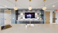 Located at 994 W Second St in Winston-Salem, NC, this single-floor amenity space blends contemporary design with industrial character to create a dynamic, multifunctional environment. The entrance greets visitors with a vibrant, oversized dot-patterned accent wall that transitions from teal to warm amber, setting an energetic and artistic tone from the moment of arrival. The open-plan layout is anchored by exposed brick columns that add warmth and texture, contrasting beautifully with the polished concrete floors and crisp white ceilings. A spacious lounge area features stylish upholstered seating arranged around a round coffee table beneath a sculptural brass globe chandelier, bathed in natural light from floor-to-ceiling windows and sliding glass doors that open to an outdoor terrace. Adjacent to the lounge, a large dining area accommodates a long communal table surrounded by woven wire chairs, with the bold dot mural serving as a striking backdrop. The kitchenette is outfitted with slate-toned cabinetry, a quartz waterfall island, a brass faucet, and a dramatic geometric tile feature wall framing a wall-mounted display screen, all illuminated by globe pendant lights with brass hardware. A game room area completes the space, featuring a ping pong table and an industrial-style roll-up garage door that opens the room to the outdoors, enhancing the open, loft-like atmosphere.