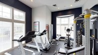This dedicated fitness space is thoughtfully designed to provide a fully functional workout environment. The room is defined by striking navy blue accent walls that contrast elegantly with crisp white upper walls and ceilings, creating a bold yet focused atmosphere ideal for exercise. The vaulted ceiling adds a sense of openness and volume to the space, preventing any feeling of confinement despite the room's concentrated purpose. Natural light floods the interior through multiple windows, including a prominent bay window that brightens the equipment area and offers views of the surrounding exterior. The flooring features durable, dark rubber matting suited for high-impact activity. A treadmill from Spirit, an elliptical trainer, a cable machine, and a weight bench are among the key pieces of equipment present, making the space suitable for both cardiovascular and strength training workouts. Recessed lighting supplements the natural light, ensuring the room is well-lit at any time of day. A fitness chart mounted on the wall serves as a practical reference for exercise routines. The layout is efficiently organized to allow comfortable movement between stations, making this a versatile and well-appointed space for fitness enthusiasts.