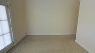 This single-floor space presents a clean and understated interior defined by its neutral design palette and practical finishes. The room visible features smooth, cream-colored walls that create a bright and open atmosphere, complemented by crisp white baseboards that provide a tidy, polished border along the floor line. The flooring consists of wall-to-wall carpet in a muted, earthy tone, offering a soft and comfortable surface underfoot. The overall aesthetic is simple and versatile, providing a blank canvas well-suited to a variety of uses and furnishing styles. The space is well-lit with an even distribution of light across the walls and floor, enhancing the sense of openness. The minimal design approach ensures that the space feels uncluttered and functional, making it highly adaptable. Standard electrical outlets are positioned along the walls, reflecting practical, everyday utility. The combination of neutral wall tones, soft carpeting, and clean architectural lines gives this space a calm, inviting ambiance that can easily be personalized to suit a range of needs and preferences.