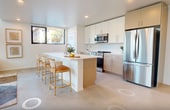 Located at The Minn in San Diego, CA, this 2-bedroom, 2-bathroom residence spans two floors and 1,150 square feet of thoughtfully designed living space. The ground floor (600 sq ft) features an open-plan living room and kitchen anchored by soaring double-height ceilings and floor-to-ceiling windows that flood the space with natural light and outdoor views. The living area is defined by polished concrete floors and a clean, minimalist aesthetic. The kitchen is outfitted with stainless steel appliances, white upper cabinetry paired with warm wood lower cabinets, a large island with bar seating, and recessed lighting. A modern staircase with a sleek metal railing connects to the upper floor (560 sq ft), where the two bedrooms are located. The 150 sq ft primary bedroom features a sliding glass door with street views and access to a bathroom with a frameless glass-enclosed shower with stone tile surround. The second bedroom is bright and spacious with polished concrete floors. A dedicated closet with built-in rods offers additional storage. The second bathroom features a vanity with modern cabinetry and clean finishes. Throughout, the unit maintains a cohesive modern design with white walls, concrete flooring, and black-framed windows.