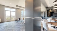This elegantly designed single-floor lounge space presents a cohesive, upscale aesthetic defined by a refined neutral palette of warm greiges, deep charcoal grays, and crisp whites. The open-concept layout is anchored by a striking coffered ceiling with recessed and decorative grid lighting that draws the eye upward, reinforcing the generous sense of volume throughout. Sleek steel-clad architectural columns divide the space while adding a contemporary industrial contrast to the soft furnishings below.

The main lounge area features plush sectional sofas and accent chairs arranged around low-profile coffee tables, all set atop patterned, multi-tone carpeting that adds texture and visual depth. A dedicated wet bar serves as a focal point, showcasing a dramatic dark marble slab backsplash with veined detailing, dark countertops, and bar seating for several guests. Adjacent to the bar, a dining and social zone includes white upholstered chairs, wood-topped tables, pendant lighting, and a large-format wall-mounted display.

Floor-to-ceiling windows and glass doors flood the space with natural light and provide direct access to an outdoor terrace, offering open cityscape views. Curated large-scale artwork punctuates the walls, contributing to the gallery-like ambiance. Every detail reflects a thoughtfully designed space intended for both social gatherings and relaxed lounging.