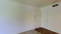 This single-floor space presents a clean, minimalist interior characterized by white-painted walls and a combination of carpet and hard flooring. The image reveals an entry area featuring a solid white front door equipped with a deadbolt and standard hardware, providing a secure main entrance. Adjacent to the entry door is a secondary door, likely leading to a closet or additional room, maintaining the same white finish for a cohesive look. The walls are painted in a crisp white throughout, contributing to a bright and open feel. The flooring transitions from dark hardwood or laminate near the entryway to carpet in the adjacent area, offering both durability and comfort underfoot. The layout appears straightforward and functional, with clean baseboards lining the walls. The overall ambiance is understated and neutral, providing a blank canvas that can be easily personalized. The space is well-suited for those seeking a simple, low-maintenance environment with practical finishes.