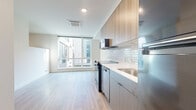 Located at 21 Boston St in Seattle's Queen Anne neighborhood, this modern 2-bedroom, 1-bathroom residence offers a thoughtfully designed single-floor layout with clean, contemporary finishes throughout. Light wood-tone flooring flows consistently across all rooms, creating a cohesive and airy atmosphere.

The kitchen is a standout feature, showcasing two-tone cabinetry with light wood uppers and darker lower cabinets, a classic white subway tile backsplash, light countertops, and a full suite of black appliances including a range, microwave, dishwasher, and refrigerator. A large floor-to-ceiling window adjacent to the kitchen fills the living area with abundant natural light and offers views of the surrounding urban streetscape.

The spacious primary bedroom is generously proportioned and features the same warm hardwood-style flooring, neutral white walls, and a sliding closet door. The secondary bedroom is bright and compact with clean walls and good natural light filtering in from clerestory-style windows above.

The bathroom is finished with full white subway tile surrounding a soaking tub and shower combo, complemented by matte black fixtures and hardware, a dark wood-panel storage cabinet, and light wood flooring. A hallway connects the rooms efficiently. The unit also features recessed lighting and a cassette-style HVAC unit for year-round climate comfort.