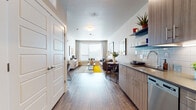 This thoughtfully designed single-floor residence offers a seamless blend of modern style and functional living. The open-concept layout connects the kitchen and living area, creating an inviting, airy atmosphere bathed in natural light from large windows dressed with layered curtains and blinds. The kitchen features flat-front wood-tone cabinetry, open shelving for display and storage, a subway tile backsplash, stainless steel appliances including a dishwasher and refrigerator, and a sleek countertop with an undermount sink. A dedicated workspace with a built-in desk area adds versatility to the floor plan. The living room is a comfortable gathering space anchored by a large window, warm hardwood-style flooring, and room for a full seating arrangement. The bedroom is generously proportioned with plush carpeting, natural light from a corner window, and ample space for a full furniture suite. The bathroom showcases a wide vanity with wood-grain cabinetry, quartz-style countertops, an undermount sink, a large mirror, and a tub-shower combination. Throughout the residence, neutral tones, clean lines, and consistent finishes create a cohesive and contemporary aesthetic.