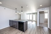 This single-floor unit showcases a clean, contemporary design with a thoughtfully arranged open-concept layout. The kitchen serves as a central hub, anchored by a large island with a granite-style countertop, an integrated sink, and a built-in dishwasher. Pendant lights with mason jar-style fixtures hang above the island, adding a modern touch. Dark-toned cabinetry lines the kitchen walls, complemented by black appliances including a refrigerator, range, and microwave. Light hardwood-style flooring flows throughout the main living areas, creating a cohesive and airy feel.

The living area receives abundant natural light through large windows and a set of French doors that open to an outdoor space, further brightening the interior. Exposed ductwork adds an industrial accent to the otherwise neutral palette of white and light gray walls.

The bedrooms feature plush carpeting and individual windows that invite natural light. A bathroom includes a walk-in shower with a built-in bench and a vanity topped with a stone-look countertop. A dedicated laundry area with in-unit washer and dryer hookups adds everyday convenience. Overall, the unit balances functionality with modern, understated aesthetics.
