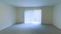 This two-story space offers a clean, functional layout designed to maximize comfort and usability across both levels. The living area, visible in the provided image, is finished with wall-to-wall carpeting in a neutral tone that complements the light-colored walls, creating a calm and cohesive atmosphere throughout. A prominent feature of this room is the large sliding glass door dressed with vertical blinds, which serves as both a source of abundant natural light and a direct connection to an outdoor area. When the blinds are drawn, sunlight floods the interior, brightening the entire space. The smooth, white textured ceiling adds to the airy feel of the room. The neutral palette throughout provides a versatile backdrop, making it easy to personalize with various furnishings and décor styles. The second floor extends the living space further, offering additional room configurations suited for a variety of needs. Overall, the space maintains a straightforward, well-lit character with practical finishes and a layout that supports everyday living across two floors.