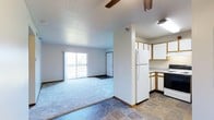 This single-floor space showcases an open-concept design that seamlessly connects the kitchen and living area. The kitchen is equipped with white cabinetry accented by warm wood-toned trim, a full-size white refrigerator, and dark appliances, including a range. The flooring in the kitchen features durable tile in a stone-like pattern, providing a practical and clean aesthetic. A structural column divides the kitchen from the living area, adding architectural definition to the open layout. The living area transitions to plush carpeting, creating a comfortable and inviting atmosphere. A textured ceiling runs throughout the space, adding subtle visual interest overhead. Ceiling fans are installed in both areas, ensuring efficient air circulation. The living room is anchored by a sliding glass door that opens to a private balcony, allowing natural light to filter into the space and offering an outdoor retreat. A secondary door is also visible, providing additional access. The neutral color palette of whites and light grays throughout creates a clean, versatile backdrop suitable for a variety of interior styles.