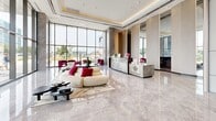 Located at 265 Boulevard Brunswick in Pointe-Claire, Quebec, this impressively scaled lobby and amenities floor spans over 6,200 square feet of thoughtfully designed communal space. The grand entrance lobby immediately captures attention with its polished marble floors, soaring ceilings adorned with a sculptural chandelier, and floor-to-ceiling glass walls that flood the space with natural light and offer open views of the surrounding greenery. Warm gold-toned cove lighting and refined stone columns add to the sense of luxury throughout.

The amenities include two distinct living areas, each with its own character. The first is a sophisticated lounge featuring rich hardwood flooring, bold fuchsia velvet accent chairs, circular pendant lighting, and curated artwork — ideal for social gatherings or relaxed lounging. A dedicated screening room outfitted with warm walnut built-ins, leather seating, and a large projection screen offers a more private retreat. A second, more casual lounge area provides additional seating with a wall-mounted display and glass-partitioned rooms beyond.

Throughout, the design balances contemporary elegance with comfort, combining high-gloss marble, natural wood finishes, designer furnishings, and generous proportions to create a welcoming and refined shared environment for residents.
