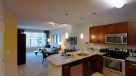This single-floor residence showcases a thoughtfully designed open-concept layout that seamlessly connects the kitchen and living areas, creating a cohesive and functional living environment. The kitchen is a standout feature, outfitted with rich, warm-toned wood cabinetry, granite countertops, and a curved peninsula that serves as both a workspace and casual dining surface. A stainless steel sink, dishwasher, and range complement the modern aesthetic, while pendant lighting above the peninsula adds an elegant touch. Track lighting overhead provides adjustable illumination throughout the space.

The open layout flows naturally into the dining and living areas, where neutral-toned carpeting adds warmth underfoot. Large windows along the far wall invite generous natural light, brightening the entire space and offering views of the surrounding urban environment. The living area is spacious enough to comfortably accommodate seating and supplementary furniture arrangements. Recessed lighting and floor lamps contribute to a layered lighting scheme. The overall ambiance strikes a balance between contemporary style and everyday comfort, making this a well-appointed and practical living space.