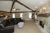This single-floor space is defined by its striking vaulted ceilings adorned with dark wood exposed beams, creating a dramatic yet warm architectural focal point throughout. A ceiling fan anchors the center of the living area, complementing the soaring ceiling height and enhancing the open, airy atmosphere.

The living room serves as the heart of the space, highlighted by a floor-to-ceiling stacked stone fireplace with a wood mantel, adding a rustic, inviting character. Large windows along the walls allow generous natural light to fill the room, brightening the neutral taupe walls and mixed-texture carpeted flooring.

The open-concept layout flows seamlessly from the living area into a transition zone featuring tile flooring, leading to a bright dining area with additional windows and ample space for a full dining set. A partial column and low partition subtly define each zone while preserving the expansive, connected feel of the interior.

Throughout the space, warm neutral tones, carpeted and tiled flooring combinations, and abundant natural light contribute to a cohesive, comfortable, and welcoming ambiance suited for everyday living and entertaining alike.