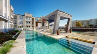 Located at 900 Grange Hall Dr in Euless, Texas, this resort-style outdoor pool area is a standout amenity designed for relaxation and entertainment. The centerpiece is a generously sized swimming pool with crystal-clear turquoise water, framed by a travertine-tiled deck that invites lounging under the Texas sun. A striking modern cabana serves as the focal point of the space, constructed with bold architectural columns and a wood-paneled ceiling, housing ceiling fans and recessed lighting for comfortable use day and night. Beneath the cabana, outdoor seating areas provide a shaded retreat overlooking the pool. A tiered water feature adds visual interest along one edge of the pool, creating a serene, resort-like ambiance. Rows of blue chaise lounges line the expansive deck, offering ample space for sunbathing. An outdoor kitchen equipped with built-in grills and prep surfaces makes the area ideal for gatherings. The surrounding landscape features ornamental grasses, drought-tolerant plantings, and raised garden beds bordered by white masonry walls. The multi-story residential buildings surrounding the pool area complete the enclosed, community-centered atmosphere of this well-appointed outdoor space.