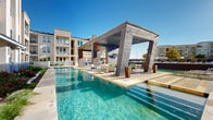 Located at 900 Grange Hall Dr in Euless, Texas, this resort-style outdoor pool area is a standout amenity designed for relaxation and entertainment. The centerpiece is a generously sized swimming pool with crystal-clear turquoise water, framed by a travertine-tiled deck that invites lounging under the Texas sun. A striking modern cabana serves as the focal point of the space, constructed with bold architectural columns and a wood-paneled ceiling, housing ceiling fans and recessed lighting for comfortable use day and night. Beneath the cabana, outdoor seating areas provide a shaded retreat overlooking the pool. A tiered water feature adds visual interest along one edge of the pool, creating a serene, resort-like ambiance. Rows of blue chaise lounges line the expansive deck, offering ample space for sunbathing. An outdoor kitchen equipped with built-in grills and prep surfaces makes the area ideal for gatherings. The surrounding landscape features ornamental grasses, drought-tolerant plantings, and raised garden beds bordered by white masonry walls. The multi-story residential buildings surrounding the pool area complete the enclosed, community-centered atmosphere of this well-appointed outdoor space.