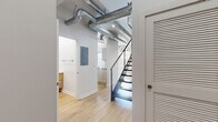 Located at 232 19th St NW in Atlanta, GA, this two-story loft blends industrial character with modern finishes throughout. The main level offers a generous open-concept living and kitchen area bathed in natural light from large multi-pane windows. The kitchen is outfitted with stainless steel appliances, sleek flat-front cabinetry, and dark granite countertops that extend to a breakfast bar. Exposed concrete ceiling beams and visible ductwork lend an authentic industrial aesthetic, while light hardwood flooring flows throughout both levels, creating warmth and continuity. A striking staircase with a modern cable railing leads to the upper level, where an arched triple-window wall floods the space with natural light and urban views. The spacious primary bedroom features soaring ceilings, a modern pendant light fixture, and access to a well-appointed walk-in closet with wire shelving. Four bathrooms serve the home, including one with a wall-mounted wood-toned vanity and round mirror, and another featuring a deep soaking tub. A dedicated utility room and additional storage complete the functional layout of this distinctive Atlanta loft.