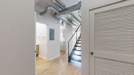 Located at 232 19th St NW in Atlanta, GA, this two-story loft blends industrial character with modern finishes throughout. The main level offers a generous open-concept living and kitchen area bathed in natural light from large multi-pane windows. The kitchen is outfitted with stainless steel appliances, sleek flat-front cabinetry, and dark granite countertops that extend to a breakfast bar. Exposed concrete ceiling beams and visible ductwork lend an authentic industrial aesthetic, while light hardwood flooring flows throughout both levels, creating warmth and continuity. A striking staircase with a modern cable railing leads to the upper level, where an arched triple-window wall floods the space with natural light and urban views. The spacious primary bedroom features soaring ceilings, a modern pendant light fixture, and access to a well-appointed walk-in closet with wire shelving. Four bathrooms serve the home, including one with a wall-mounted wood-toned vanity and round mirror, and another featuring a deep soaking tub. A dedicated utility room and additional storage complete the functional layout of this distinctive Atlanta loft.