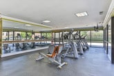 This fitness center in Phenix City, Alabama offers a comprehensive workout facility designed to accommodate a wide range of exercise routines. The space is finished with durable rubber flooring throughout, providing a safe and supportive surface for both cardio and strength training activities.

The cardio zone features a row of treadmills and elliptical machines positioned along expansive floor-to-ceiling windows, flooding the space with natural light and offering open views of the surrounding outdoor area. A wall-mounted television is positioned above the cardio equipment for entertainment during workouts.

The strength training area is well-appointed with a rack of dumbbells and weight plates, along with multiple cable and resistance machines suited for a full-body workout. An exercise chart is displayed on the wall, serving as a helpful reference for users. A dedicated stretch and functional training zone includes resistance bands and additional cable equipment.

Large mirrored wall panels enhance the sense of space and allow for form monitoring during exercise. The interior is finished with bold yellow-green accent walls trimmed with crisp white crown molding, giving the space a clean and energetic aesthetic. Recessed and flush-mounted lighting supplements the abundant natural light. The single-floor layout ensures easy accessibility, making this fitness center a practical and inviting space for daily use.