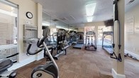 Located at 4008 Gaelic Ln in Glen Allen, Virginia, this dedicated fitness center offers a well-equipped, single-floor space designed to support a comprehensive workout routine. The room features warm, neutral-toned walls paired with patterned carpet flooring, creating a functional yet comfortable environment for exercise.

The cardio section is generously outfitted with multiple treadmills, elliptical trainers, and stationary and recumbent bikes, providing ample options for aerobic training. A strength training area includes a multi-station cable machine and free weights, offering a full range of resistance workout options. Large floor-to-ceiling mirrors line one wall, amplifying the sense of space and allowing users to monitor their form during exercise.

Natural light flows in through several large windows fitted with blinds, offering views of the surrounding outdoor community. The bright, overhead fluorescent lighting ensures the space remains well-lit at all times. A wall-mounted clock and a water fountain add practical conveniences, while posted fitness center rules near the entry door reinforce a structured, community-oriented environment. The space is accessible via a dedicated exterior door, making it easy to enter directly from the surrounding grounds.