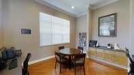 This single-story space presents a welcoming and functional layout highlighted by warm hardwood flooring that runs throughout the main area. The room is finished with elegant crown molding along the ceiling, adding a refined architectural detail that elevates the overall aesthetic. A large window fitted with white horizontal blinds allows ample natural light to fill the space, creating a bright and comfortable atmosphere.

At the center of the room sits a round dark wood dining table accompanied by four matching dark wood chairs with cushioned seats, providing a practical and inviting setting for small group gatherings or casual meetings. Along one wall, a wall-mounted display rack holds an assortment of reading materials or informational pamphlets, suggesting the space may serve a hospitality or community purpose.

To the right, light wood cabinetry with a dark countertop supports a coffee station, complete with a coffee maker and condiment organizer, further reinforcing the space's function as a communal lounge or hospitality area. A framed mirror mounted on the adjacent wall adds depth and reflects additional light into the room. The neutral taupe wall tones tie all elements together cohesively.