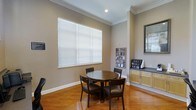 This single-story space presents a welcoming and functional layout highlighted by warm hardwood flooring that runs throughout the main area. The room is finished with elegant crown molding along the ceiling, adding a refined architectural detail that elevates the overall aesthetic. A large window fitted with white horizontal blinds allows ample natural light to fill the space, creating a bright and comfortable atmosphere.

At the center of the room sits a round dark wood dining table accompanied by four matching dark wood chairs with cushioned seats, providing a practical and inviting setting for small group gatherings or casual meetings. Along one wall, a wall-mounted display rack holds an assortment of reading materials or informational pamphlets, suggesting the space may serve a hospitality or community purpose.

To the right, light wood cabinetry with a dark countertop supports a coffee station, complete with a coffee maker and condiment organizer, further reinforcing the space's function as a communal lounge or hospitality area. A framed mirror mounted on the adjacent wall adds depth and reflects additional light into the room. The neutral taupe wall tones tie all elements together cohesively.