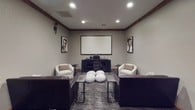This dedicated screening room delivers a refined cinematic experience within a single, thoughtfully designed space. The room is anchored by a large wall-mounted projection screen paired with a ceiling-mounted projector, supported by a dark wood media console that houses the audiovisual equipment. Warm wood beam accents line the upper perimeter of the neutral gray walls, adding an architectural detail that lends the room a sophisticated, classic character.

Seating is both plentiful and varied, combining dark brown leather sofas and armchairs with cream upholstered barrel chairs, all arranged to face the screen and encourage comfortable viewing. A cluster of chunky knit ottomans at the center of the room provides flexible additional seating and a relaxed, casual accent. Industrial-style side tables are positioned throughout for convenience.

The walls are adorned with a curated collection of classic Hollywood photography and film-themed artwork, reinforcing the cinematic theme with a refined touch. Recessed lighting illuminates the space evenly, while wall-mounted torch-style sconces contribute to the theatrical ambiance. Patterned carpet in muted tones ties the room together and enhances the acoustics. A solid wood double door provides a grand entry point, completing this well-appointed screening room.